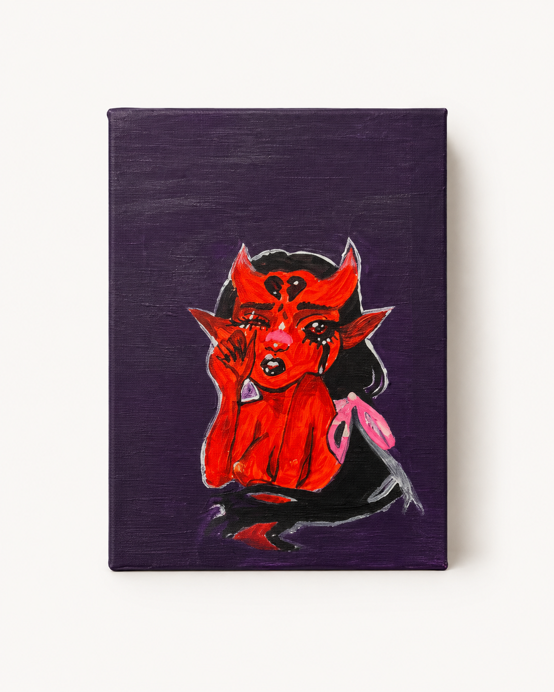 A vertical canvas art print featuring a stylized red devil character with horns and a pink bow against a dark purple background, shown on a light off-white wall.

Devil in Her Feelings original red character painting on black canvas